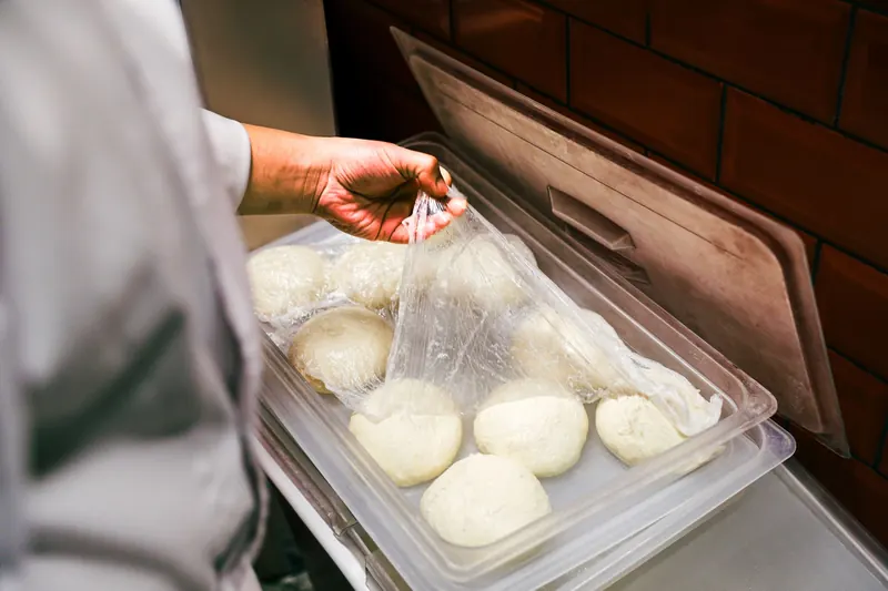 Chef lifting plastic wrap from fresh dough balls in a kitchen setting