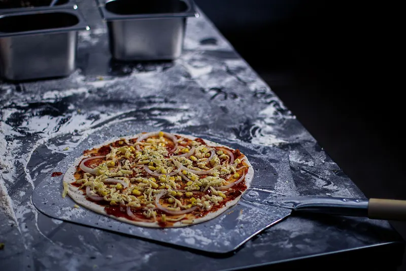 Close-up of uncooked pizza with cheese, onion, and corn on a baking peel, ready for the oven