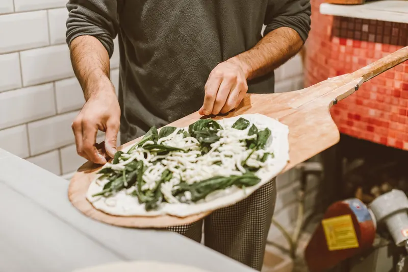 A person crafting a spinach and cheese pizza with a wooden paddle near a traditional rustic oven